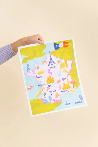 A hand holds a colorful illustrated map poster of France against a solid light beige background. The map features cartoon landmarks like the Eiffel Tower, Notre Dame, and the Louvre, with the French flag in the top right corner.