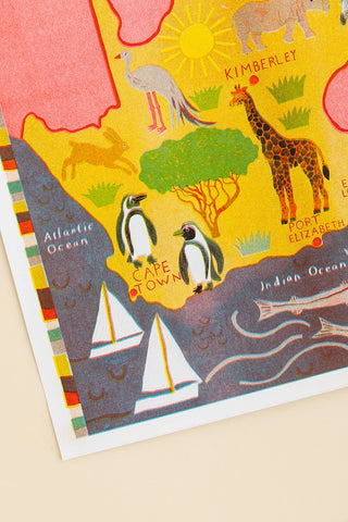 A close-up of a colorful illustrated map or poster showing parts of Africa, with labeled oceans, sailboats, penguins, a giraffe, a bird, and text including Kimberlet and Fort Elizabeth, photographed on a light beige background.