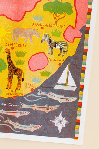 A colorful illustrated map print of South Africa showing landmarks, animals like a zebra and giraffe, cities such as Johannesburg and Durban, fish in the ocean, and a sailboat, photographed against a light neutral background.