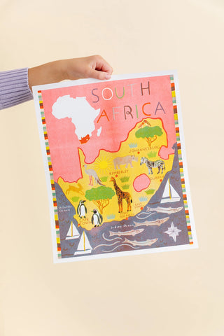 A hand holding a colorful South Africa map art print against a solid light beige background. The print features a pink landmass with a white silhouette of Africa, yellow interior details, and a dark gray ocean with illustrated penguins, a giraffe, sailboats, and a star.