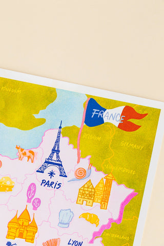 A close-up of a colorful illustrated map print featuring Paris, with the Eiffel Tower, a France flag, and other travel-themed icons on a cream background.
