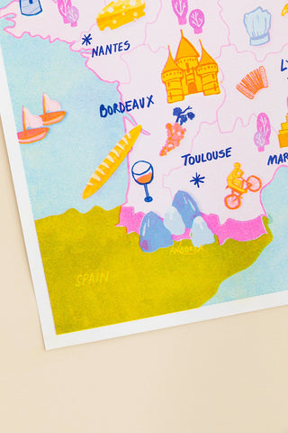 A close-up of a colorful illustrated map print featuring French cities like Nantes, Bordeaux, and Toulouse, along with small travel-themed icons, photographed on a light beige background.